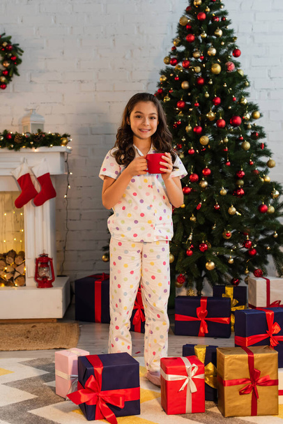 Bambino sorridente in pigiama che tiene la tazza vicino all'albero di Natale e i regali a casa  - Foto, immagini