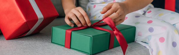 Cropped view of child in pajama untying bow on Christmas gift on couch, banner  - Foto, Imagem