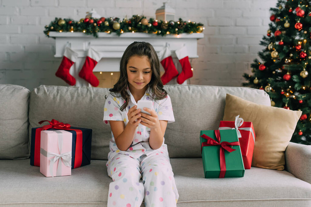 Chica en pijama utilizando el teléfono celular cerca de los regalos de Navidad y pino decorado en casa  - Foto, Imagen