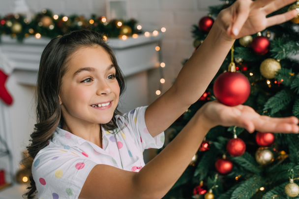 Cheerful girl in pajama holding blurred ball near Christmas tree at home  - 写真・画像