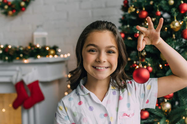 Kid in pajama holding ball near blurred Christmas tree at home  - Photo, Image