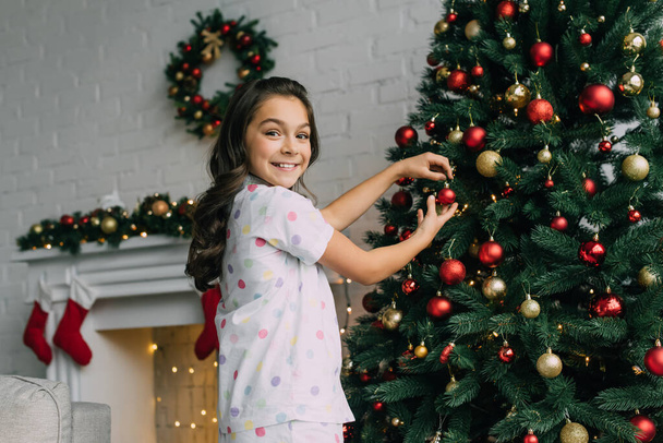Lächelndes Mädchen im Pyjama schmückt Tanne zu Weihnachten zu Hause  - Foto, Bild