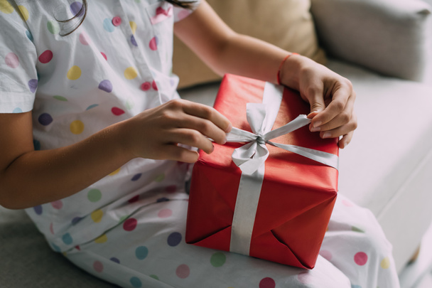 Cropped view of child in pajama holding Christmas gift with bow on couch  - Фото, изображение
