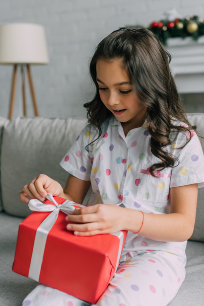Niño en pijama punteada ajustando lazo en regalo de Navidad en casa  - Foto, imagen