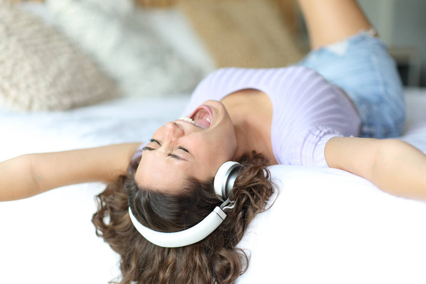 Excited woman singing and listening to music wearing headphones lying on a bed in a room - Foto, Imagem