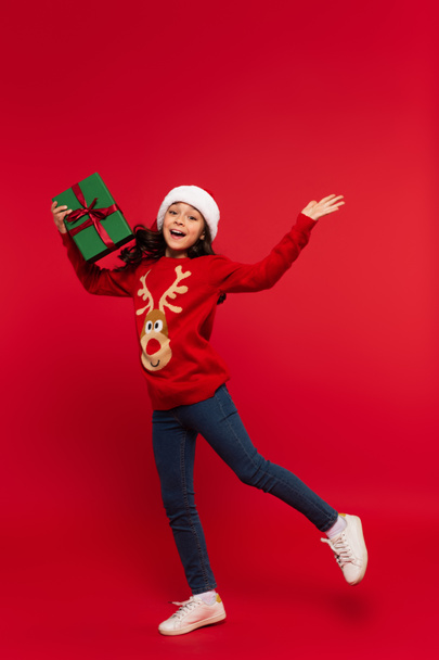 Cheerful girl in Christmas sweater and santa hat holding gift box on red background  - Valokuva, kuva