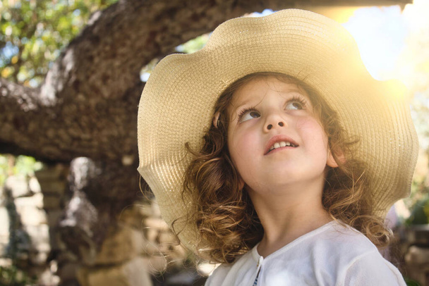 Close-up of a cute little girl wearing a rural straw hat looking up with glowing sunlight in the background - Фото, изображение