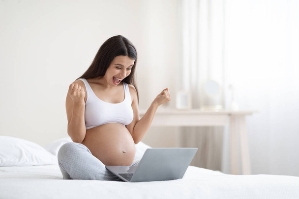 Émotionnel jeune femme enceinte aux cheveux longs dans des vêtements confortables assis sur le lit à la maison, à l'aide d'un ordinateur, regardant l'écran d'ordinateur portable, criant et serrant les poings, jouant sur Internet, célébrant le succès - Photo, image
