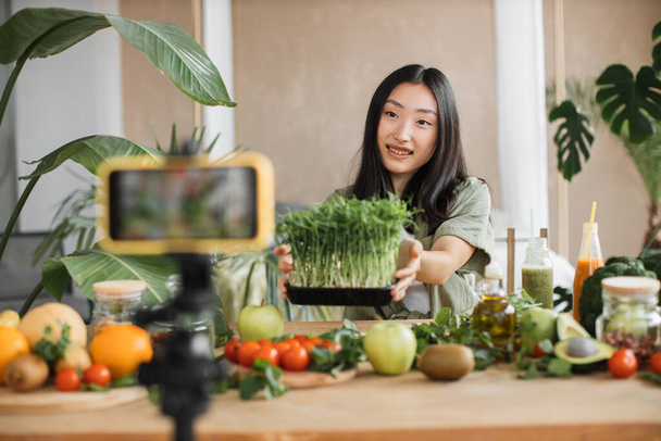 Beautiful young asian woman blogger recording video lesson on smart phone about recipe how to cook healthy salad from microgreen sprouts and organic fresh vegetables and fruits. - Φωτογραφία, εικόνα