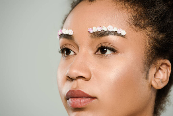 close up of pretty african american woman with flowers on eyebrows isolated on grey - Photo, image