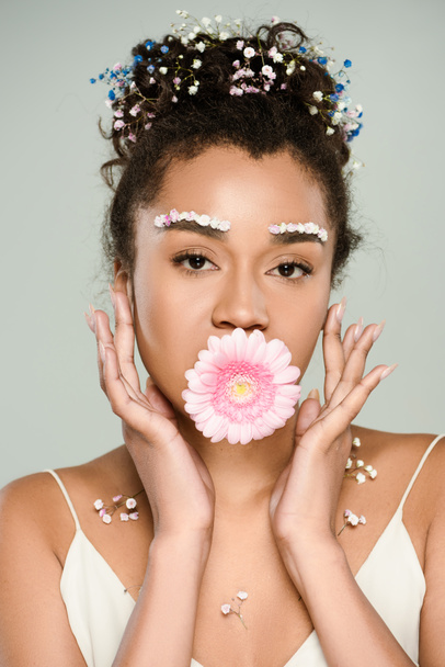 curly african american woman with flowers on eyebrows and hair with gerbera in mouth isolated on grey - Foto, immagini