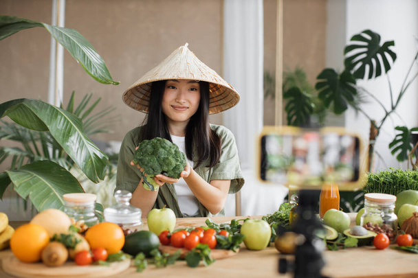 Asian woman making healthy food using phone creating video content for social media in tropical resort kitchen studio. Beautiful female using broccoli, vegetables and fruits preparing healthy salad - Foto, afbeelding
