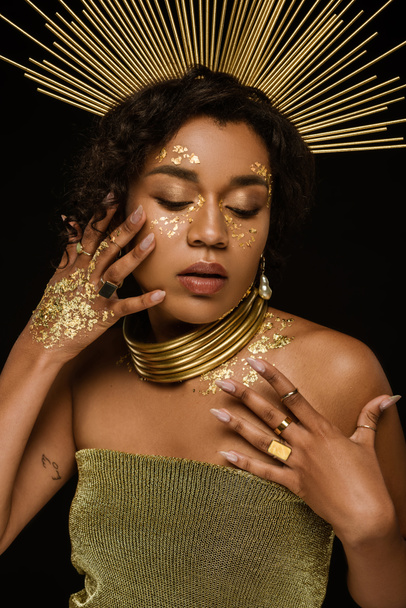 young african american woman with golden accessories and paint on face posing isolated on black - Foto, Bild