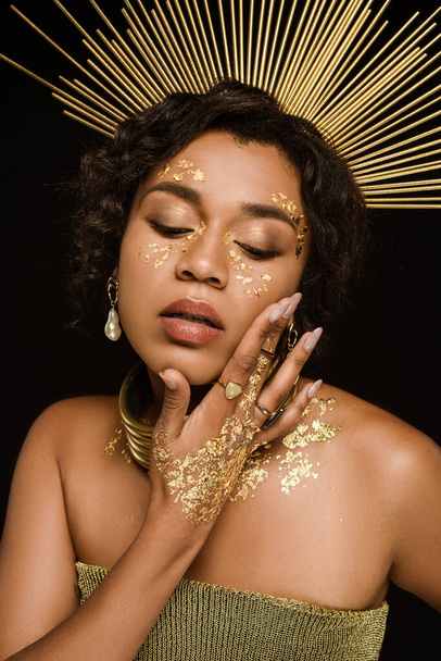 curly african american woman with golden accessories and paint on face posing isolated on black - Foto, imagen