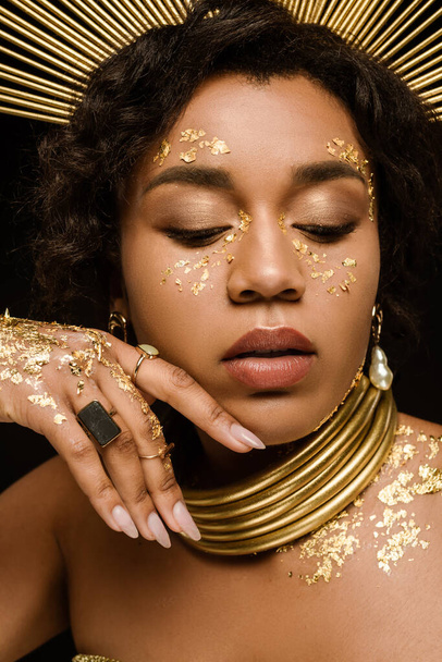 close up of african american woman with golden accessories and paint on face posing isolated on black - Foto, immagini