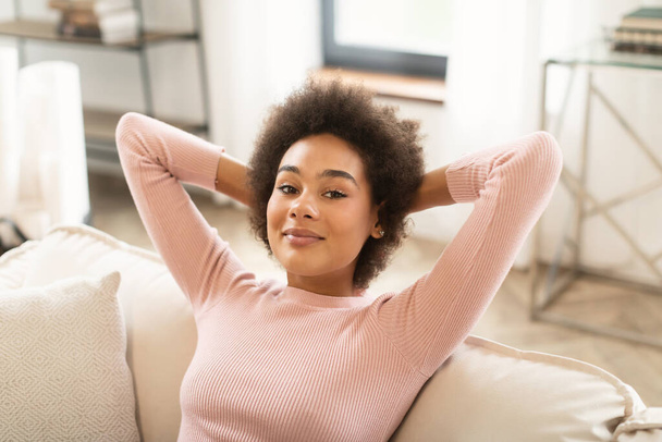 Cheerful relaxed millennial african american woman resting on sofa, enjoy comfort and cozy in bright living room interior, close up. Silence, free time and weekend, break at home alone due covid-19 - Fotografie, Obrázek