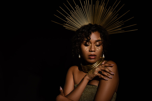 confident african american woman with golden crown and paint on cheeks posing isolated on black - Foto, imagen