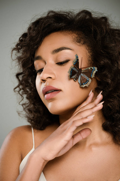 brunette african american woman with butterfly on cheek posing isolated on grey - Fotó, kép