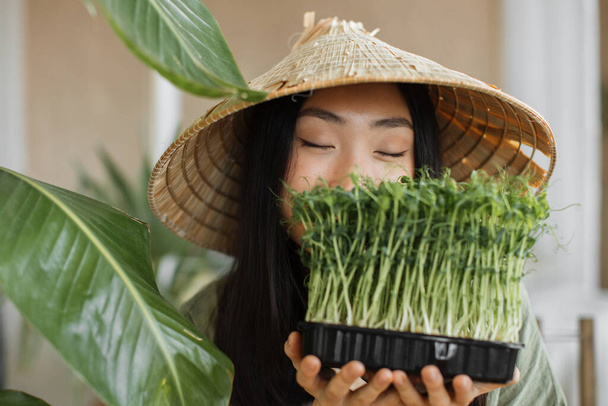 Portrait of young asian woman with closed eyes holding microgreen sprouts on tropical exotic background, enjoying healthy diet, indoor at background with palm leaves. Weight loss concept. - Foto, afbeelding