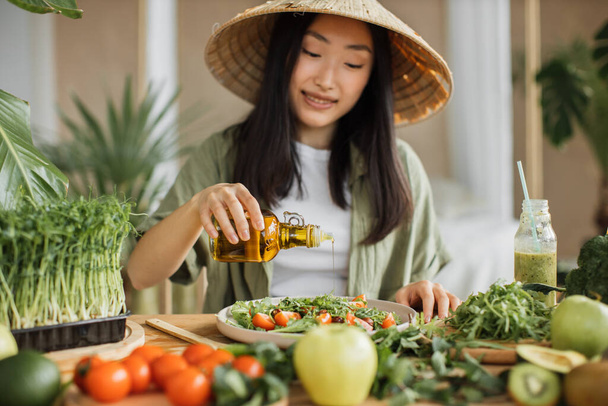 Young attractive asian woman in traditional conical hat sitting at table in stylish light tropical kitchen and pouring olive oil to healthy vegetable salad. - Zdjęcie, obraz