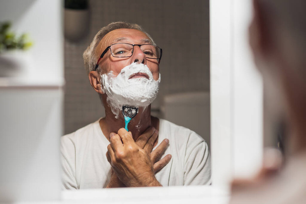 Старший мужчина бреет бороду бритвой и пены для бритья в ванной. - Фото, изображение