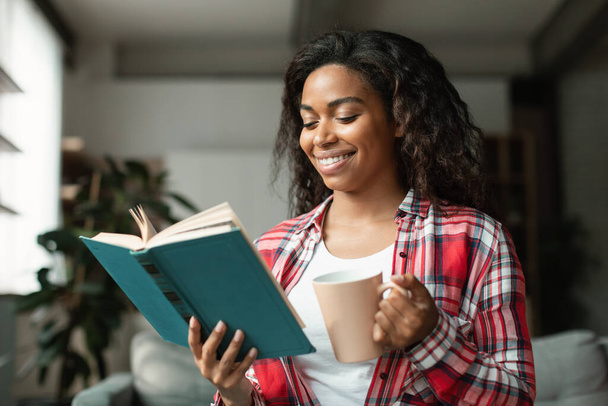Cheerful smart pretty black young woman student reading book, enjoy cup of hot favorite drink, study and spare time in living room interior. Literature, hobby, coffee break at home alone, covid-19 - Foto, Imagen