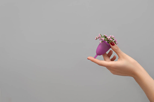 Vrouwelijke hand houden menstruele beker gevuld met bloemen op grijze achtergrond, vrouw welzijn concept  - Foto, afbeelding
