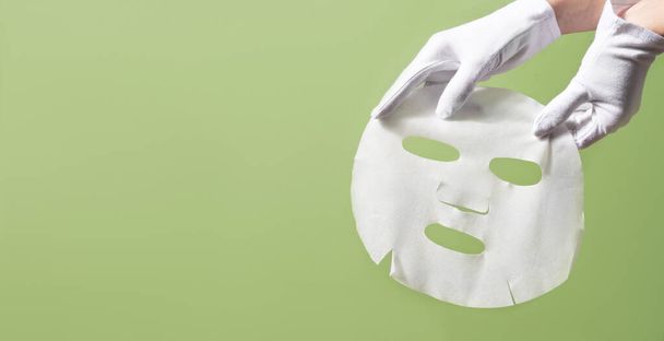 Female gloved hands holding white facial mask sheet on green background. Beauty skincare concept - Photo, Image