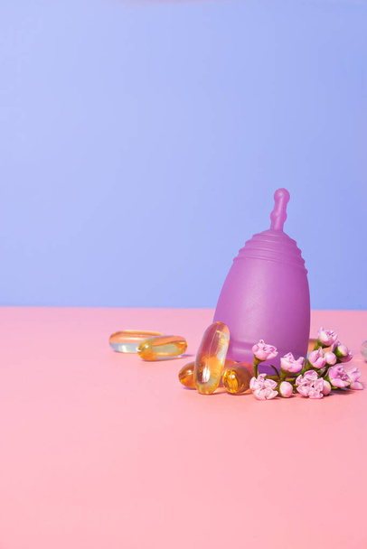 Dişi menstual bardak, çiçekler ve yağ kapsülleri pembe masada. Kadın refahı kavramı   - Fotoğraf, Görsel