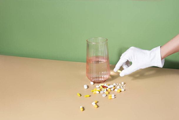 Female gloved hand holding a pill. glass of water and handful of pills are on a table. Well-being concept  - Foto, imagen