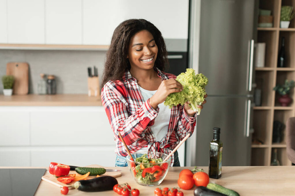 Fröhliche schwarze junge Dame in lässigem Salat, bereitet Speisen aus Bio-Gemüse zu, hat Spaß in der modernen Kücheneinrichtung. Kochen gesunde vegetarische hausgemachte essen allein zu Hause, richtige Ernährung, covid-19 - Foto, Bild