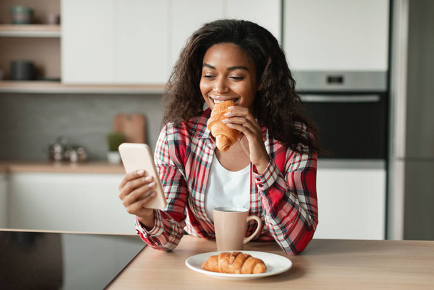 Cieszy czarna młoda kobieta w casual jeść croissant, wpisując na telefon, rozmowy, czytanie wiadomości i cieszyć się śniadaniem, dzień dobry w nowoczesnym wnętrzu kuchni. Nowa aplikacja social media, surfing w internecie w domu - Zdjęcie, obraz