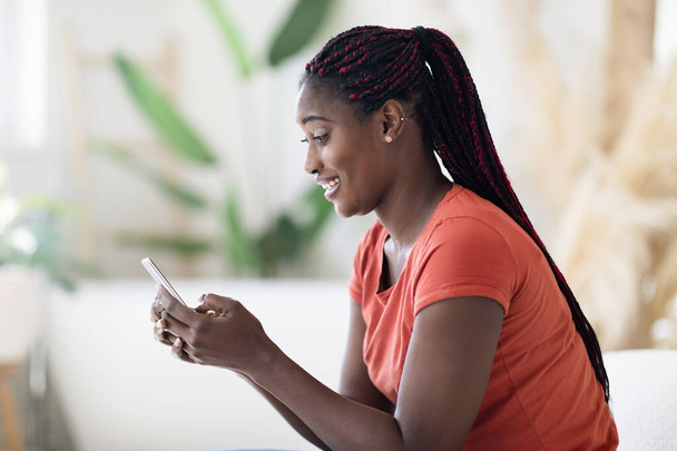 Мобільні повідомлення. Посмішка чорна жінка використовує смартфон під час відпочинку вдома, щаслива молода афроамериканська жінка СМС з друзями або браузер Інтернет На мобільному телефоні, сторонній погляд - Фото, зображення