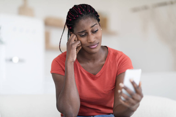 Znudzona młoda czarna kobieta patrząc na ekran smartfona siedząc na kanapie w domu, zamyślona Afroamerykanka trzymająca telefon komórkowy, czekając na ważne połączenie lub Sms, skopiuj przestrzeń - Zdjęcie, obraz