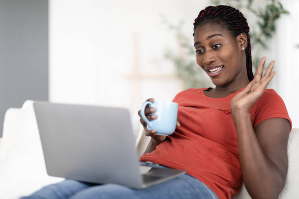 Kaukainen kommunikaatio. Onnellinen musta nainen tekee videopuhelun kannettavalla tietokoneella ja juo kahvia istuessaan sohvalla kotona, nuori afroamerikkalainen nainen, jolla on etäkonferenssi tietokoneella, lähikuva - Valokuva, kuva