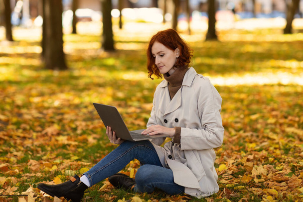 Skoncentrowana biała, tysiącletnia kobieta z czerwonymi włosami w płaszczu przeciwdeszczowym siedzi na ziemi z żółtymi liśćmi, pisząc na komputerze, surfując w internecie, studiując samotnie jesienią. Praca i biznes z gadżet na świeżym powietrzu - Zdjęcie, obraz