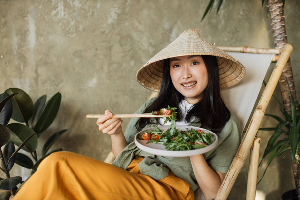 Portrait of young asian woman holding vegetarian salad while sitting on folding chair with organic vegetables, enjoying healthy diet, indoor at grey background. Weight loss concept. - Фото, изображение