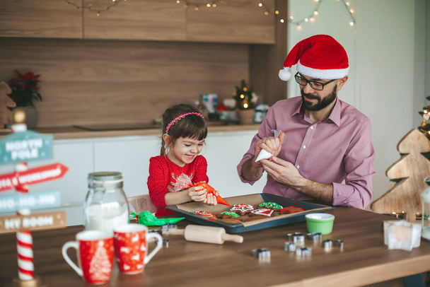 Family having fun decorating gingerbread Christmas cookies. Father and his little daughter preparing for Christmas. - Fotografie, Obrázek