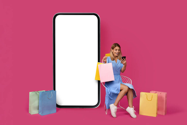 Шокированная улыбающаяся взрослая европейская женщина сидит на стуле со многими сумками и смотрит на телефон рядом с огромным смартфоном с пустым экраном, изолированным на фиолетовом фоне. Приложение для предложений и рекламы, продаж и онлайн-покупок - Фото, изображение