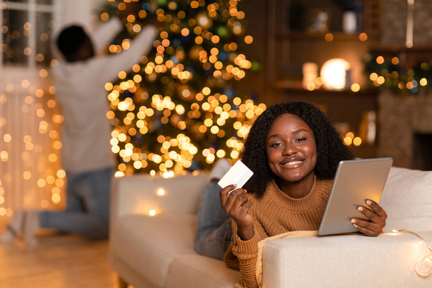 Joyeux jeune femme noire avec tablette, carte de crédit profiter des achats en ligne, vente de vacances, mari décore arbre de Noël dans le salon intérieur avec des guirlandes. Nouvel An célébration, commande app - Photo, image
