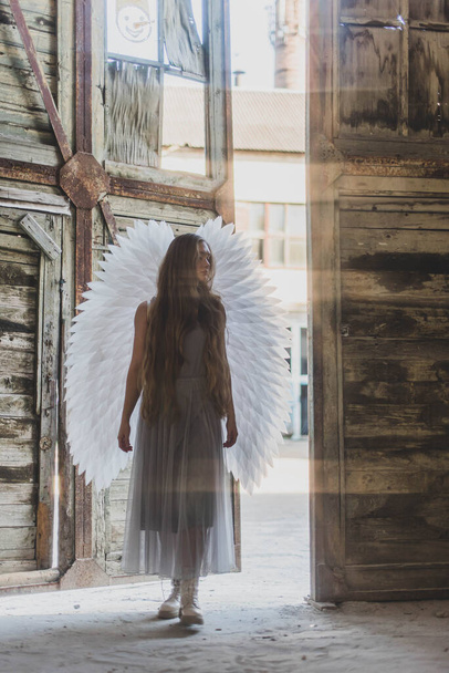 Une fille aux cheveux longs avec de grandes ailes blanches. La fille est un ange. Photo en mouvement. La fille entre dans le bâtiment, ouvrant la porte en bois - Photo, image