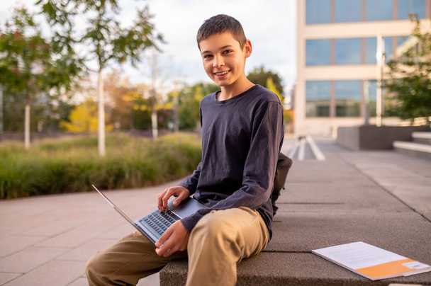 Smiling pleased teenager with the portable computer sitting on a concrete porch of a building - Fotoğraf, Görsel