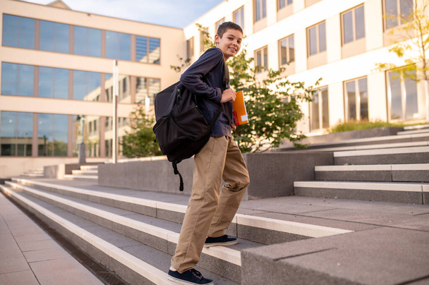 Smiling teen with the backpack over the shoulder and a textbook in the hand standing on the stairs - 写真・画像