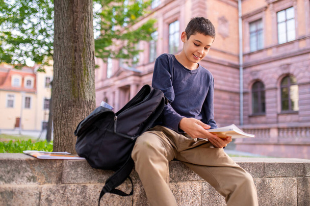 Smiling contented schoolboy sitting under a deciduous tree and looking at a textbook in his hands - Фото, изображение