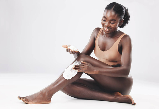 Μαύρη γυναίκα, κάθεται με κρέμα ξυρίσματος στα πόδια για περιποίηση δέρματος, καλλυντικά και ευεξία του δέρματος με χαμόγελο. Χαρούμενη Αφρικανή γυναίκα, περιποίηση μαλλιών σώματος και αυτοφροντίδα, αυτοαγάπη και υγεία από το φόντο του στούντιο. - Φωτογραφία, εικόνα