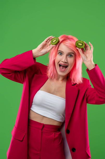 Merry extravagant fashionable girl with halves of kiwi posing for the camera against the green background - Foto, Bild