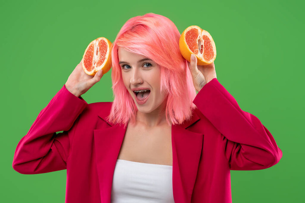 Portrait of a mischievous girl with a tongue piercing and braces holding two halves of grapefruit in her hands - Foto, Imagen