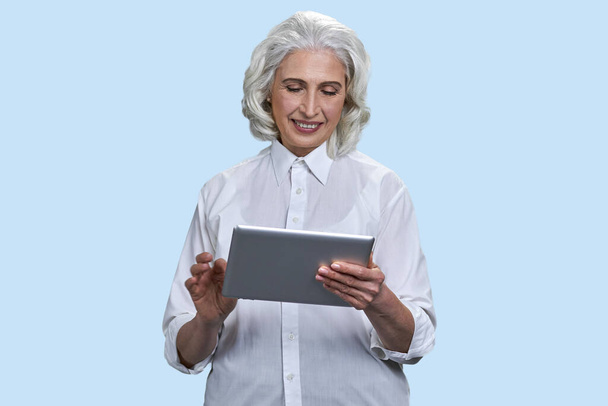 Usmívající se starší šedovlasá žena používající tablet. Izolované na světle modrém pozadí. - Fotografie, Obrázek