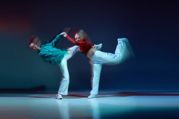 Portret van jonge man en vrouw dansend hiphop geïsoleerd over donkerblauwe achtergrond met gemengd licht. Optreden. Begrip beweging, jongerencultuur, actieve levensstijl, actie, straatdans, reclame - Foto, afbeelding