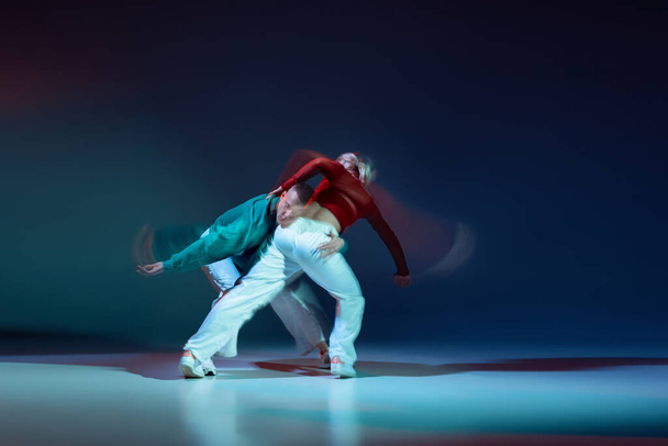 Portrait of young man and woman dancing isolated over dark blue background with mixed lights. Emotional dance. Concept of movement, youth culture, active lifestyle, action, street dance, ad - Photo, Image